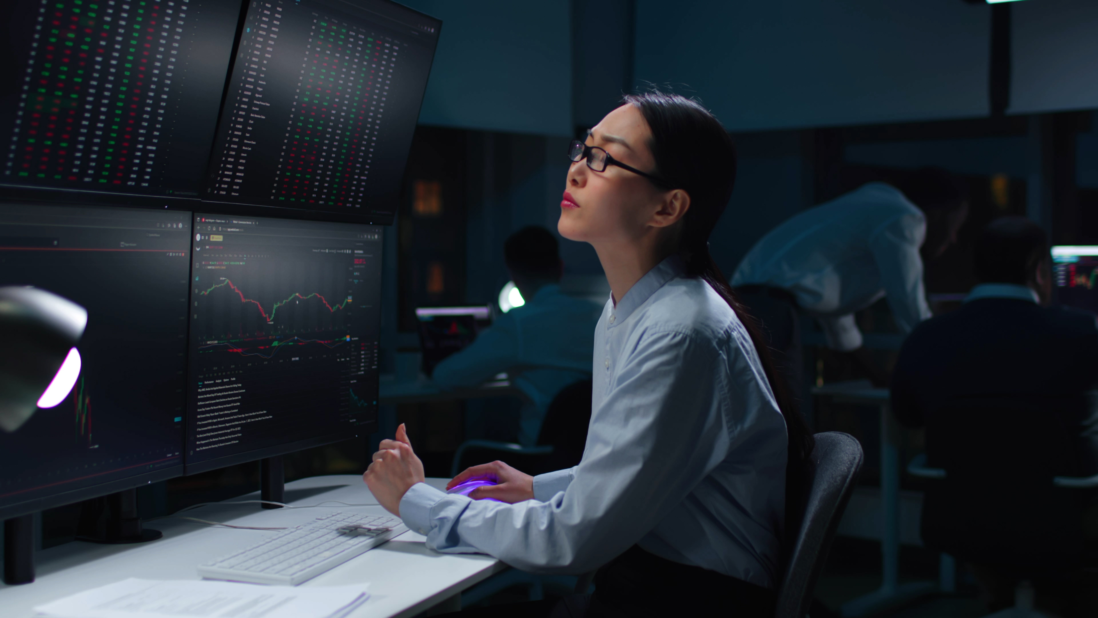 Asian woman trader look at multi-screen workstation with graphs and data working late in dark office.